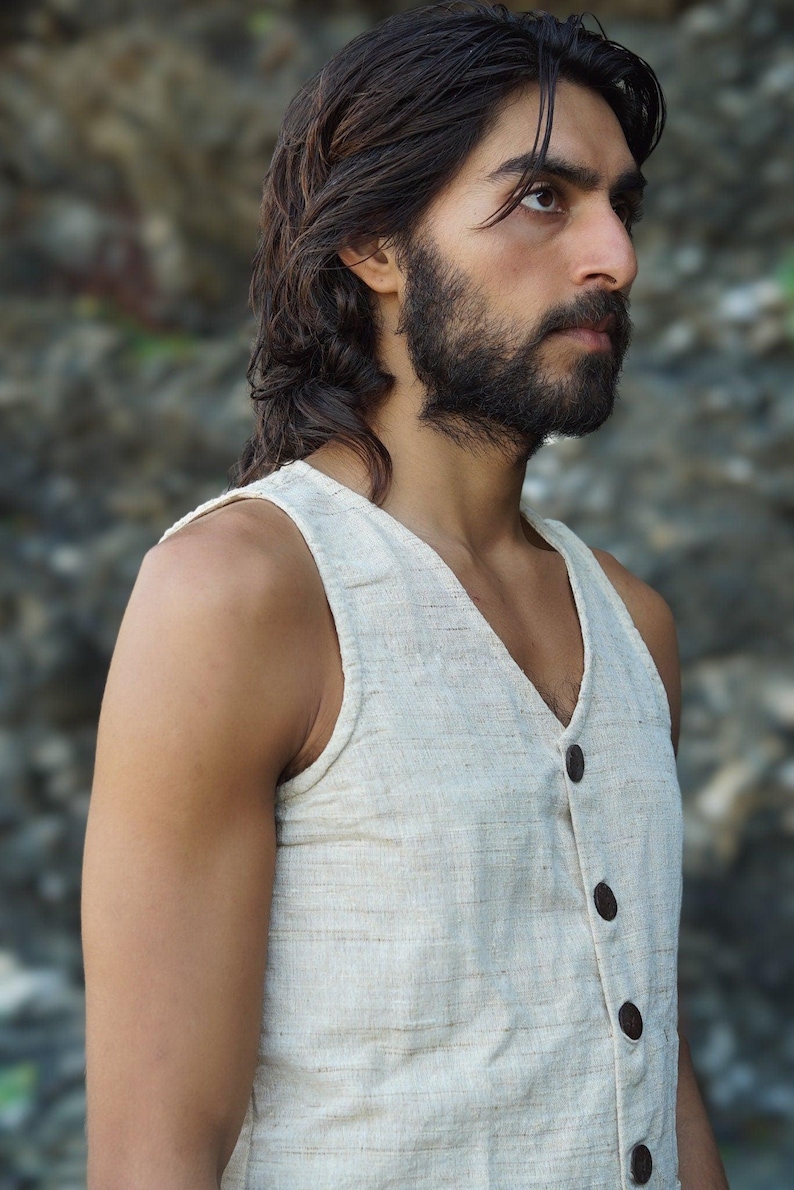 May include: A man with long dark hair and a beard is wearing a sleeveless, off-white linen vest with brown buttons. The vest has a V-neck and is made of a textured fabric.