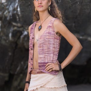 May include: A woman wearing a sleeveless, pink and white striped vest with a brown belt and a beige, layered, fringed skirt. She is wearing a necklace with a blue stone pendant and has a nose ring.