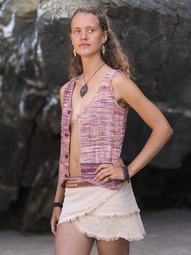 May include: A woman wearing a sleeveless, pink and white striped vest with a brown belt and a beige, layered, fringed skirt. She is wearing a necklace with a blue stone pendant and has a nose ring.