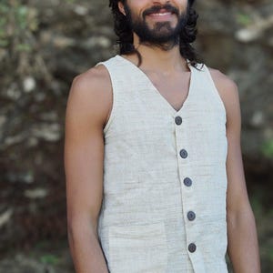 May include: A man wearing a light beige sleeveless vest with brown buttons. The vest has a pocket on the front. The man is wearing brown pants.