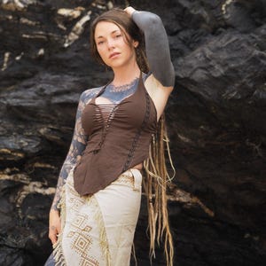 Könnte beinhalten: Eine Frau mit langen blonden Dreadlocks trägt ein braunes Halstuch mit Schnürung vorne und einen weißen Rock mit geometrischem Muster. Sie hat schwarze Tattoos auf ihren Armen und steht vor einem felsigen Hintergrund.