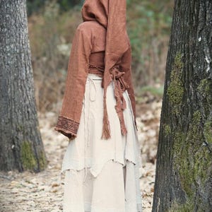 May include: A woman wearing a brown hooded top with a white tiered skirt. The top has a long, flowing sleeve and a cinched waist. The skirt is made of a lightweight fabric and has a frayed hem.