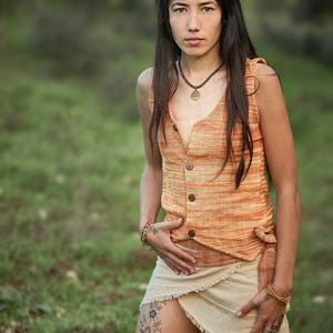 May include: A woman with long dark hair wearing a sleeveless orange and white striped top with a button-up front and a cream-colored wrap skirt. She is wearing a necklace with a circular pendant and bracelets on both wrists.