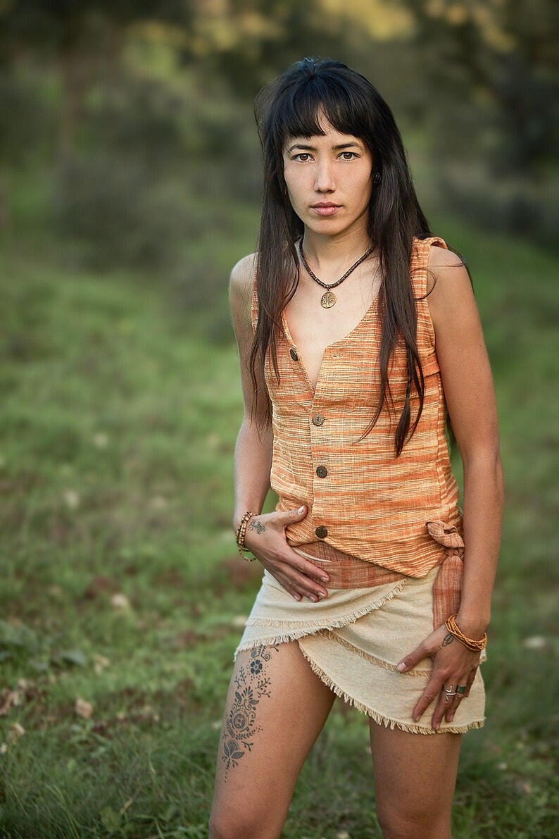 May include: A woman with long dark hair wearing a sleeveless orange and white striped top with a button-up front and a cream-colored wrap skirt. She is wearing a necklace with a circular pendant and bracelets on both wrists.