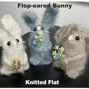 May include: Three knitted bunny figurines with floppy ears. The bunnies are holding small bouquets of flowers. The bunnies are white, light blue, and light brown. The text "Flop-eared Bunny" and "Knitted Flat" are visible in the image.