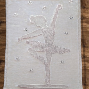 May include: A white knitted baby blanket featuring a ballerina design. The ballerina is wearing a white dress with a pink outline. The blanket is decorated with small white dots.