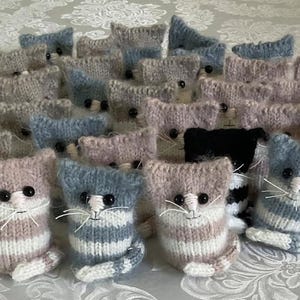 May include: A collection of knitted cat figurines in various colors, including blue, gray, pink, and black. Each cat has a striped pattern, button eyes, and white whiskers. The cats are arranged on a patterned surface.