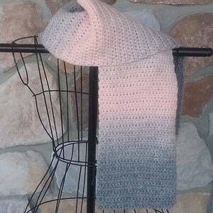 May include: A handmade crochet scarf in a gradient of colors, transitioning from light pink to gray. The scarf is displayed on a black metal form against a stone background. The scarf's texture is visible, showcasing the individual stitches.