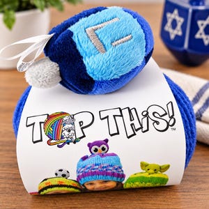 May include: A plush blue and light blue dreidel toy with a silver Hebrew letter attached to a ball of blue yarn. The yarn is wrapped in a white paper label featuring the text TOP THIS! and illustrations of children wearing hats with various plush toppers.