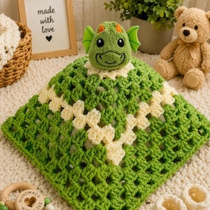 May include: A green and cream-colored crochet security blanket featuring a plush dragon head at the center. The blanket is constructed with a square granny stitch pattern in alternating shades of green and cream yarn, topped with a soft dragon head.