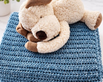 May include: A plush dog toy with cream-colored textured fur and brown accents on the ears, paws, and nose, resting on top of a folded, deep blue crocheted blanket with a consistent horizontal stitch pattern throughout the fabric.