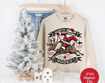 Pode incluir: Uma camisa de moletom bege com um gráfico de Papai Noel montando um rena. O gráfico inclui o texto "North Pole Rodeo" e "North Pole".