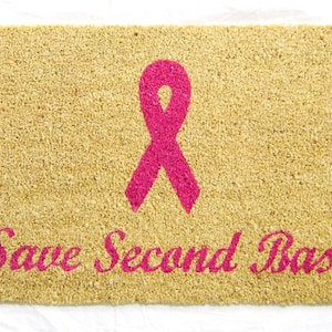 May include: A tan doormat with a pink ribbon and the text "Save Second Base" in pink.