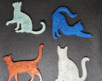 Puede incluir: Cuatro adornos con forma de gato en varios colores. Los gatos están en diferentes poses: sentados, estirándose y caminando. Los colores incluyen azul claro, azul, naranja y blanco. Los artículos parecen estar hechos de un material texturizado.