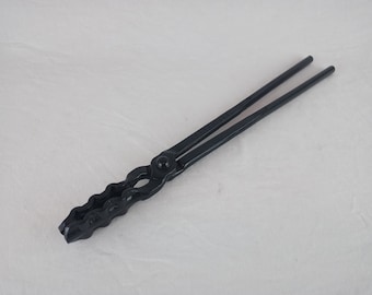May include: Black metal blacksmith tongs with a wavy gripping end and long, straight handles. The tongs are joined at a central pivot point. The tool is designed for holding hot metal during forging or other metalworking processes.