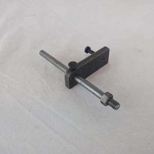 Puede incluir: Una herramienta de metal con una varilla larga y una base rectangular. La varilla tiene una tuerca y un tornillo en un extremo y una perilla en el otro extremo. La base tiene un tornillo en un lado.