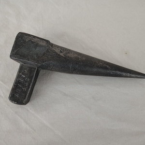 Puede incluir: Una herramienta en forma de cuña de metal oscuro, con un mango rectangular y una punta larga y afilada. El mango tiene el texto "S.M.F. CANADA" estampado. La herramienta parece ser de hierro o acero.