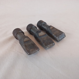 Könnte beinhalten: Drei Meißel aus Metall mit einem abgerundeten Kopf und einer flachen, spitzen Klinge. Die Meißel sind aus dunklem Metall und haben ein abgenutztes, rustikales Aussehen.