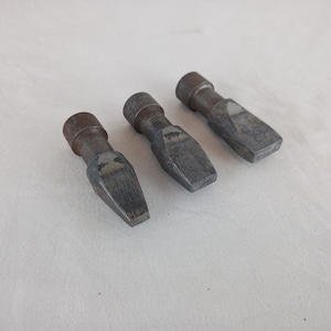 Pode incluir: Três cinzéis de metal com ponta pontiaguda e cabo arredondado. Os cinzéis são cinzentos e têm um aspeto desgastado e enferrujado.