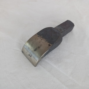 May include: A metal tool with a curved, beveled edge and a rectangular handle. The tool is made of a dark metal with a slightly rusted finish.