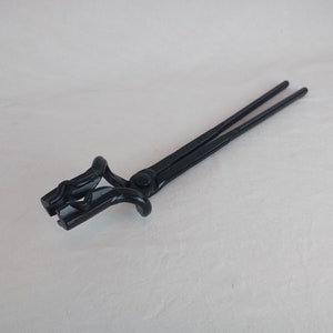 Puede incluir: Tenazas de herrero de metal negro con un diseño de mandíbula curvada. Las tenazas se utilizan para sujetar y manipular metal caliente.