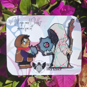 May include: Enamel pin set depicting a child and a robed mage. The child has a tear, and the mage holds a staff. The pins are on a white card with the text "Mage and Child enamel pin set" and "Witch Workshop".