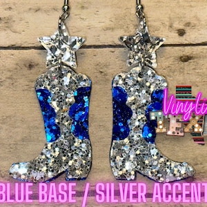 May include: A pair of blue and silver glitter boot earrings with a silver star charm. The earrings are on a wooden background with the text "Vinyltize Texas".