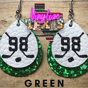 Pode incluir: Brincos de couro branco e glitter verde com o número 98 e tacos de hóquei impressos.