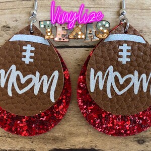 May include: A pair of brown leather earrings with red glitter accents. The earrings are shaped like footballs and have the words "# MOM" written on them in white.