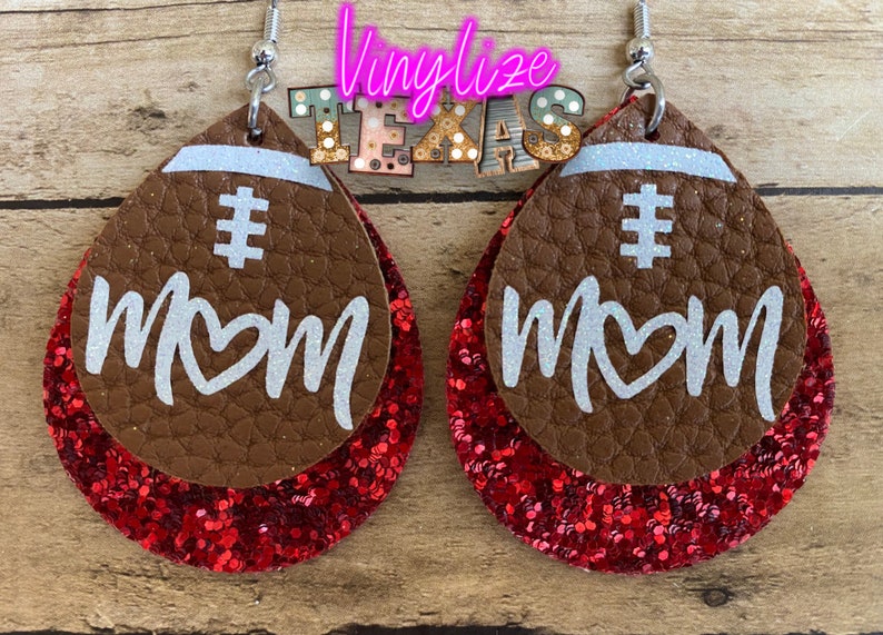 May include: A pair of brown leather earrings with red glitter accents. The earrings are shaped like footballs and have the words "# MOM" written on them in white.