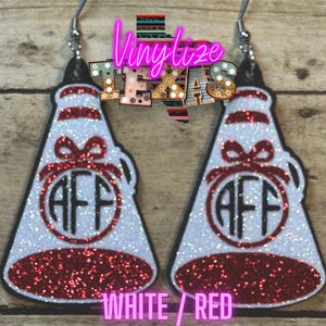 May include: A pair of white and red glitter cheerleader megaphone earrings. Each earring features a white body with red glitter accents and a black outline. The earrings have the letters "AFF" in a circle and the text "WHITE / RED".