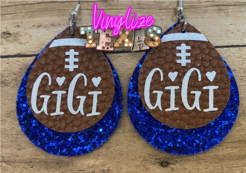May include: A pair of blue and brown glitter football-shaped earrings with the word "GIGI" and a hashtag on each earring.