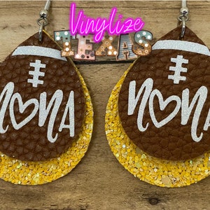 May include: A pair of brown leather earrings shaped like footballs with gold glitter accents. The earrings have the word "#MOMA" written on them in white glitter.