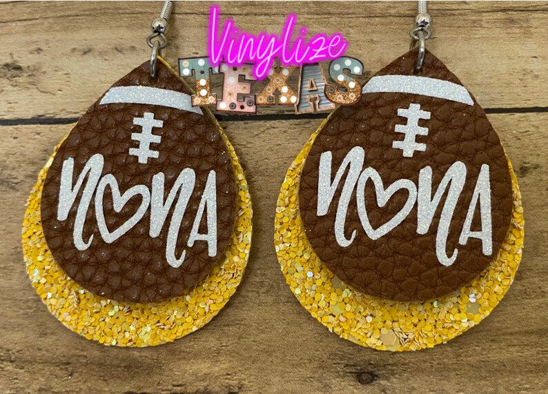 May include: A pair of brown leather earrings shaped like footballs with gold glitter accents. The earrings have the word "#MOMA" written on them in white glitter.