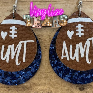 May include: A pair of brown leather earrings shaped like footballs with blue glitter accents. The earrings have white glitter text that says "# AUNT" with a white heart above the hashtag.