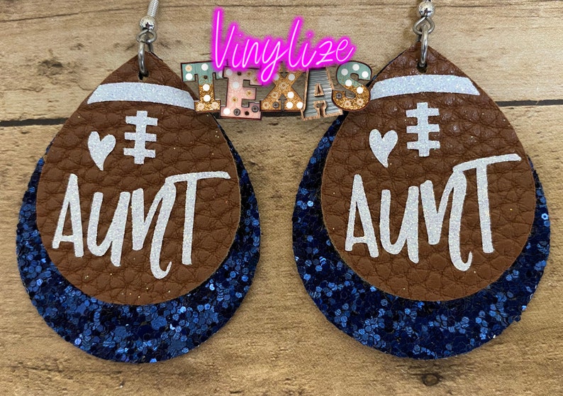 May include: A pair of brown leather earrings shaped like footballs with blue glitter accents. The earrings have white glitter text that says "# AUNT" with a white heart above the hashtag.
