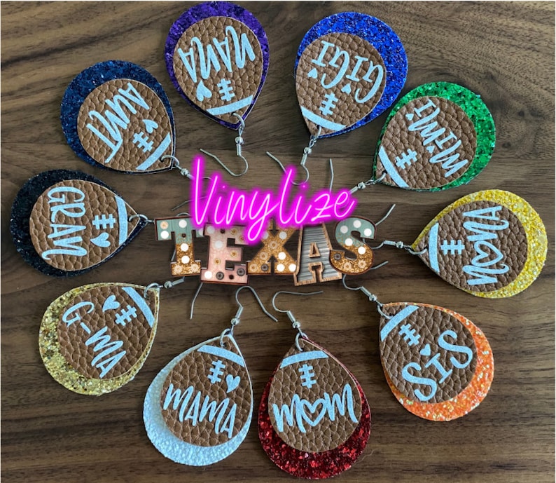 May include: A set of 10 pairs of football-shaped earrings with different glitter colors and names like "Mama", "Grandma", "Sis", and "Mom". The earrings are made of brown leather and have a white glitter finish. The earrings are displayed on a wooden surface with a sign that says "Vinylze Texas".