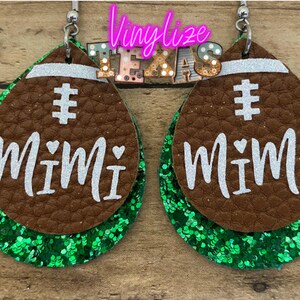 May include: A pair of brown leather earrings with green glitter accents. The earrings are shaped like footballs and have the word "Mimi" written on them in white.