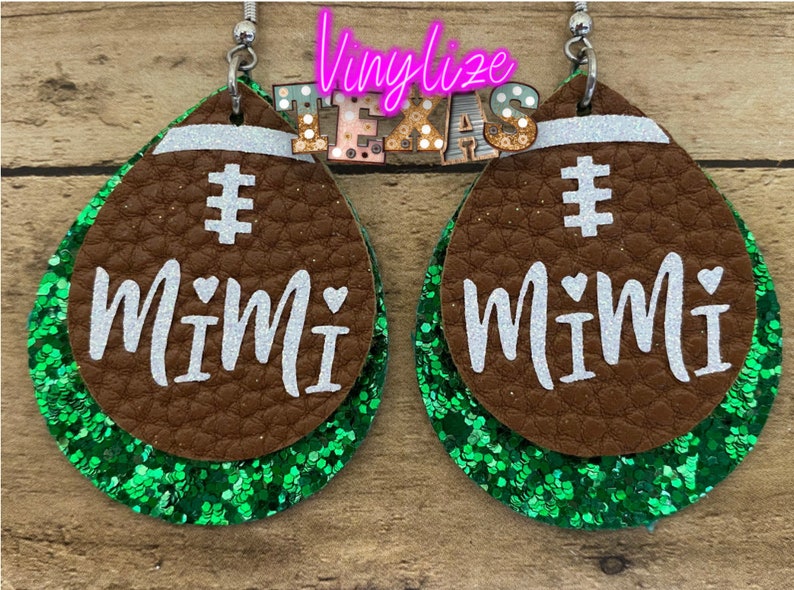 May include: A pair of brown leather earrings with green glitter accents. The earrings are shaped like footballs and have the word "Mimi" written on them in white.