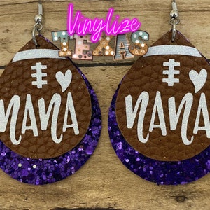May include: A pair of brown leather earrings with purple glitter accents. The earrings are shaped like footballs and have the words "# Mama" written on them in white.