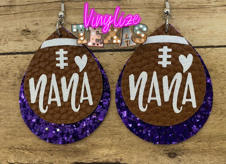 May include: A pair of brown leather earrings with purple glitter accents. The earrings are shaped like footballs and have the words "# Mama" written on them in white.