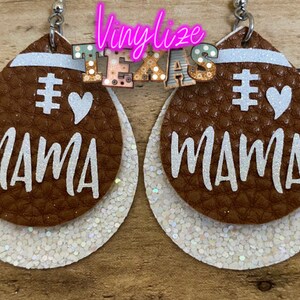 May include: A pair of brown leather earrings with white glitter accents. The earrings are shaped like footballs and have the words "#1 Mama" written on them.