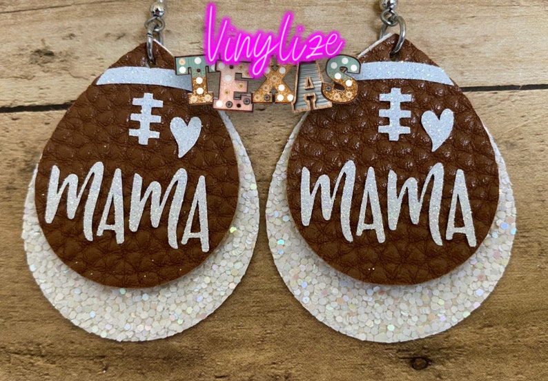 May include: A pair of brown leather earrings with white glitter accents. The earrings are shaped like footballs and have the words "#1 Mama" written on them.