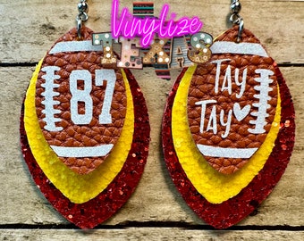 Op de afbeelding: Lederen oorbellen in de vorm van een voetbal met een gelaagd ontwerp. De oorbellen hebben bruine, gele en rode glitterlagen. Eén oorbel heeft het nummer "87" en de andere zegt "Tay Tay" met een hart. Het woord "Texas" staat bovenaan.