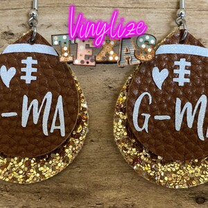 May include: A pair of brown leather earrings shaped like footballs with gold glitter accents. The earrings feature white text that says "# G-MA" and a white heart.