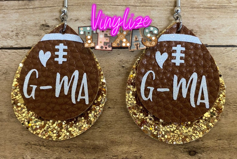 May include: A pair of brown leather earrings shaped like footballs with gold glitter accents. The earrings feature white text that says "# G-MA" and a white heart.