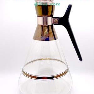 May include: A clear glass pitcher with a gold band around the middle and a black handle. The top of the pitcher is made of brass with a copper finish.