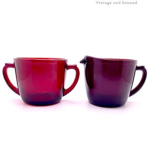 May include: Two vintage ruby red glass pieces: a mug with two handles and a creamer with a spout. The mug is on the left, and the creamer is on the right. The text "Dots Connection Vintage and beyond" is in the upper right corner.