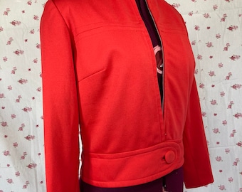 Puede incluir: Una chaqueta de color naranja brillante con cierre de cremallera y un solo botón en la parte delantera. La chaqueta tiene una cintura ajustada y mangas largas.