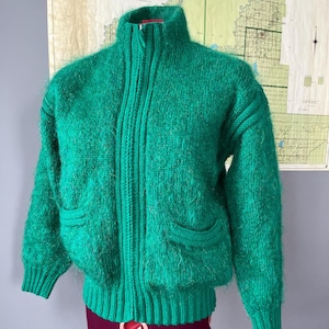 Puede incluir: Un cárdigan verde peludo con cremallera y dos bolsillos. El cárdigan está en un maniquí.